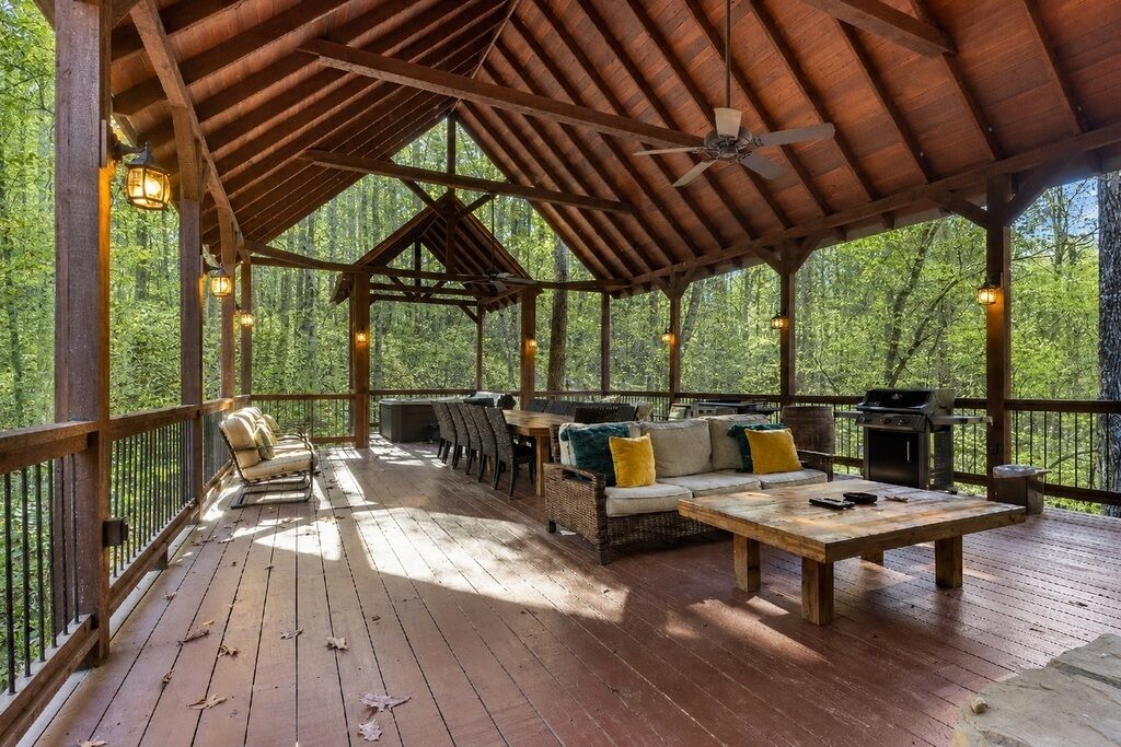 A covered outdoor deck with wooden furniture, a ceiling fan, and a view of a forest.