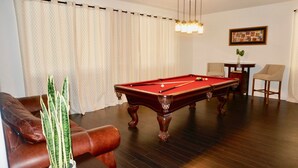 Uma mesa de bilhar em um quarto com um sofá de couro marrom, uma planta em vaso e uma mesa com cadeiras.