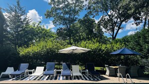 A poolside area with lounge chairs and umbrellas, surrounded by trees and a clear sky.