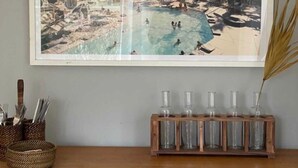 A framed photograph of a pool area with palm trees and mountains in the background, placed above a wooden cabinet with glassware.