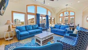 A spacious living room with large windows, blue sofas, a coffee table, and a chandelier. - Semi-Oceanfront Dog Friendly w/ Pool, Hot Tub, Elevator, Theater, and Game Room (Salvo)