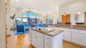 A spacious kitchen with white cabinets, a granite countertop, and a large island. There is a dining area with blue furniture and a view of the ocean through large windows. - Semi-Oceanfront Dog Friendly w/ Pool, Hot Tub, Elevator, Theater, and Game Room (Salvo)
