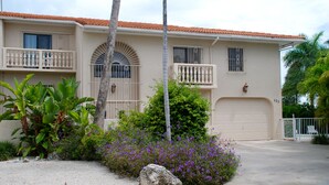 Una casa de dos pisos con garaje, rodeada de plantas tropicales y un camino de grava.