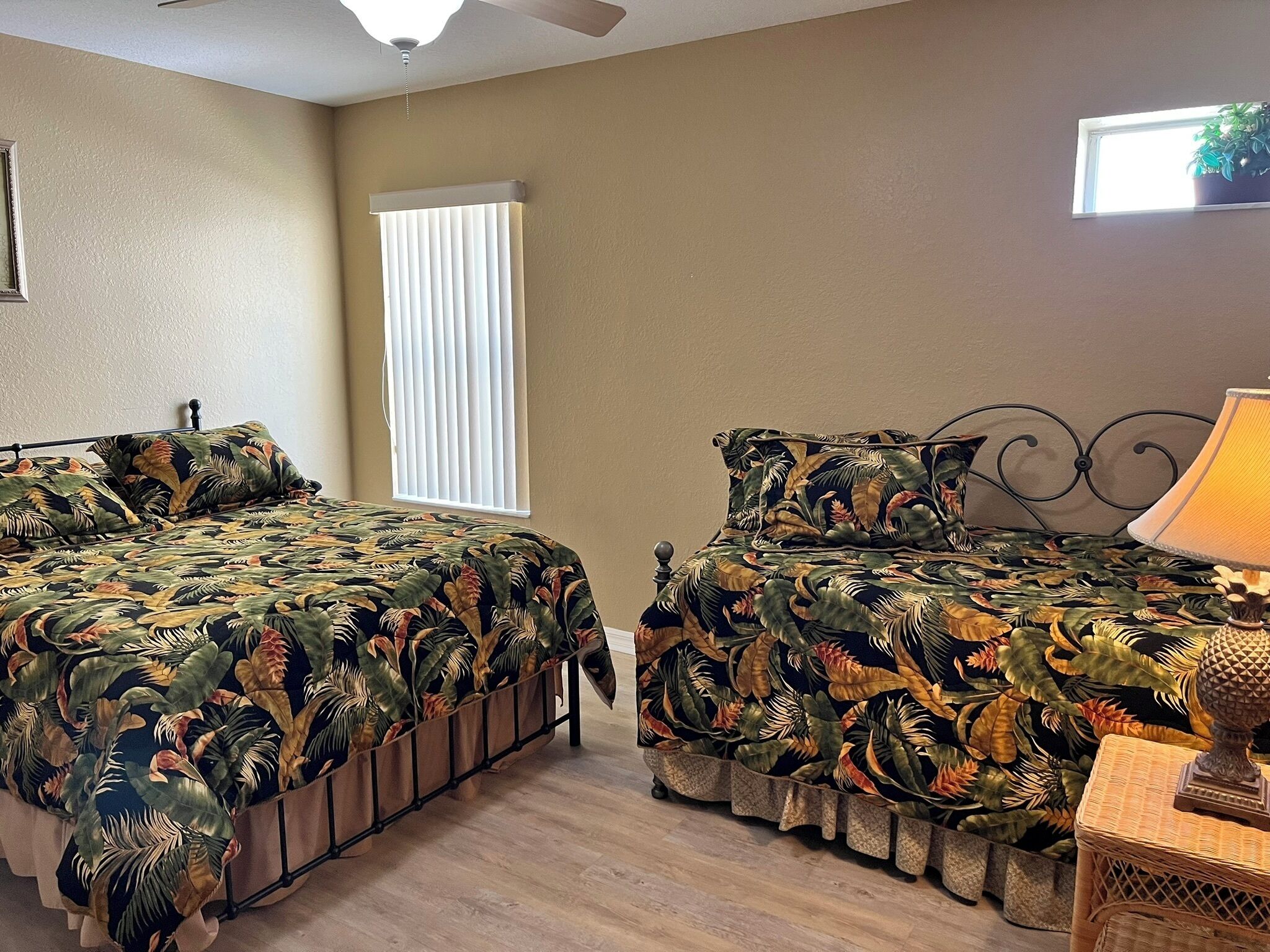 A bedroom with two beds, a ceiling fan, a window with blinds, and a bedside lamp.
