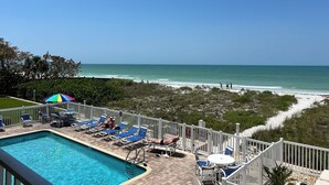 Un área de piscina con sillones de descanso y sombrillas, vista a la playa y una cerca blanca.