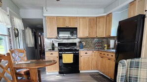 A kitchen with wooden cabinets, a black stove, a microwave, and a refrigerator. There is a dining table with chairs and a plaid sofa. - Seasonal Fully Furnished Mobile home (Port Austin)