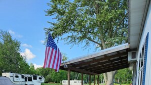 A covered patio area with outdoor seating, a flag, and a recreational vehicle in the background. - Seasonal Fully Furnished Mobile home (Port Austin)