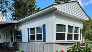 A single-story house with a white exterior and blue shutters, a gravel driveway, and a garden with various plants and flowers. - Seasonal Fully Furnished Mobile home (Port Austin)