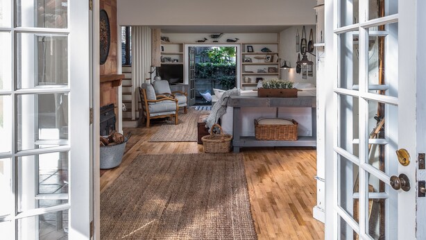 A spacious living area with a wooden floor, a sofa, a coffee table, and a dining table. There is a kitchen area with a stove and a sink visible.