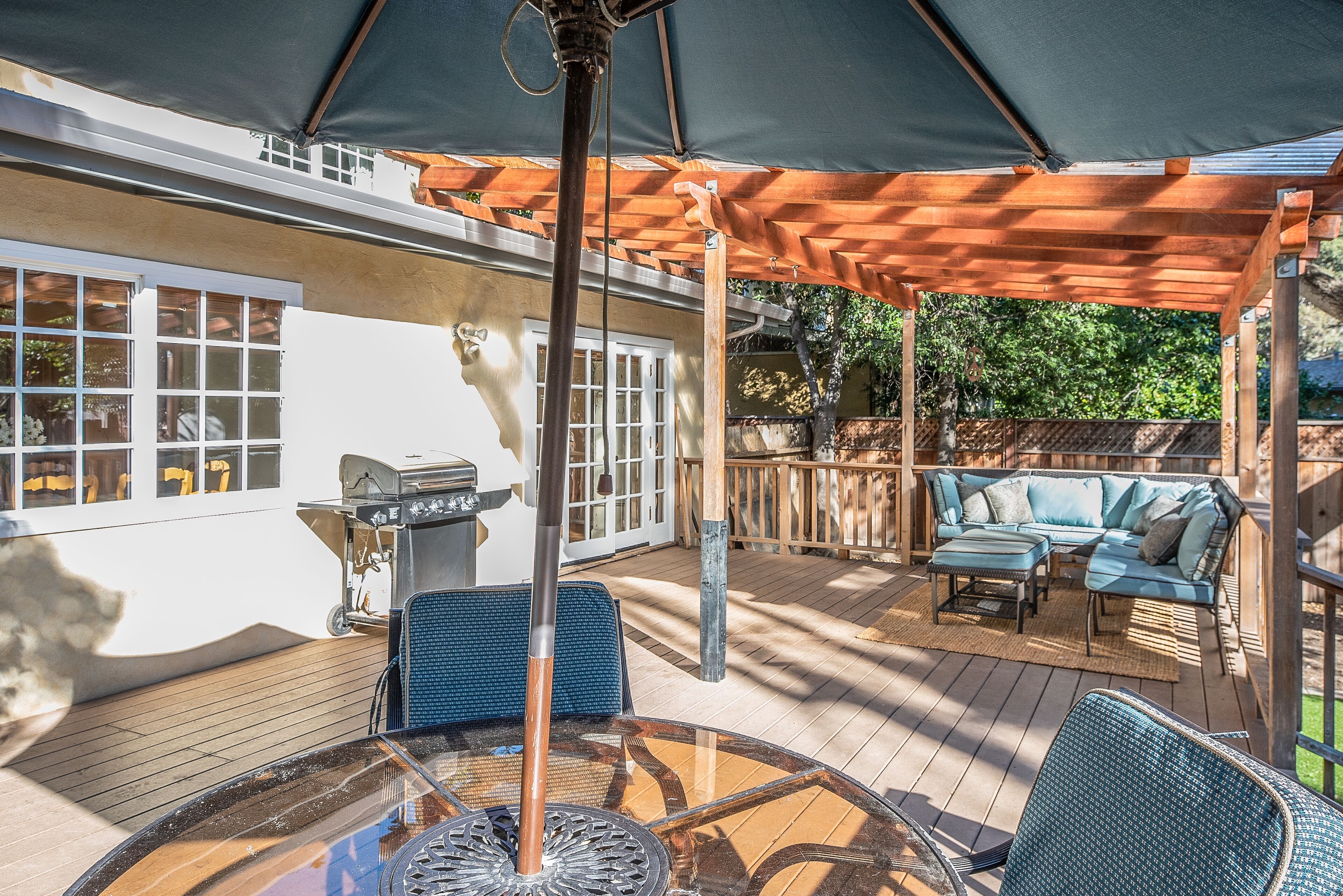 An outdoor patio with a wooden deck, a glass table, blue chairs, a barbecue grill, and a covered seating area.