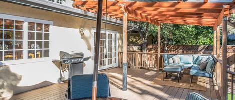 An outdoor patio with a wooden deck, a glass table, blue chairs, a barbecue grill, and a covered seating area.