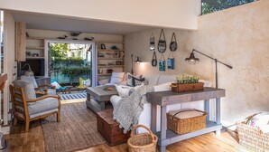 A cozy living room with a sofa, armchair, and coffee table, a wooden cabinet with decorative items, and a large window overlooking a garden.