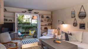 A living room with a sofa, armchairs, a coffee table, and a TV. There are shelves with decorative items and a view of the outside.
