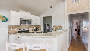 A kitchen with white cabinets, a microwave, a stove, and a coffee maker. There is a breakfast bar with two chairs. - Book now for Fall 2026! Pet friendly! (Gulf Shores)