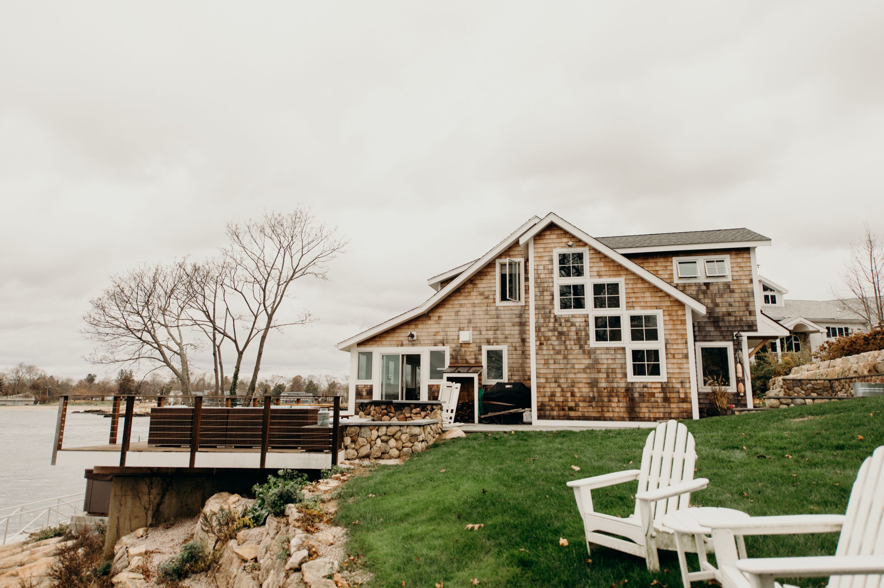 Une grande maison dotée d’une terrasse donnant sur un plan d’eau, avec un mur en pierre et une pelouse agrémentée de deux chaises Adirondack blanches.