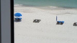 A beach view with a clear sky, calm sea, and a few people swimming. There are beach chairs and umbrellas visible.
