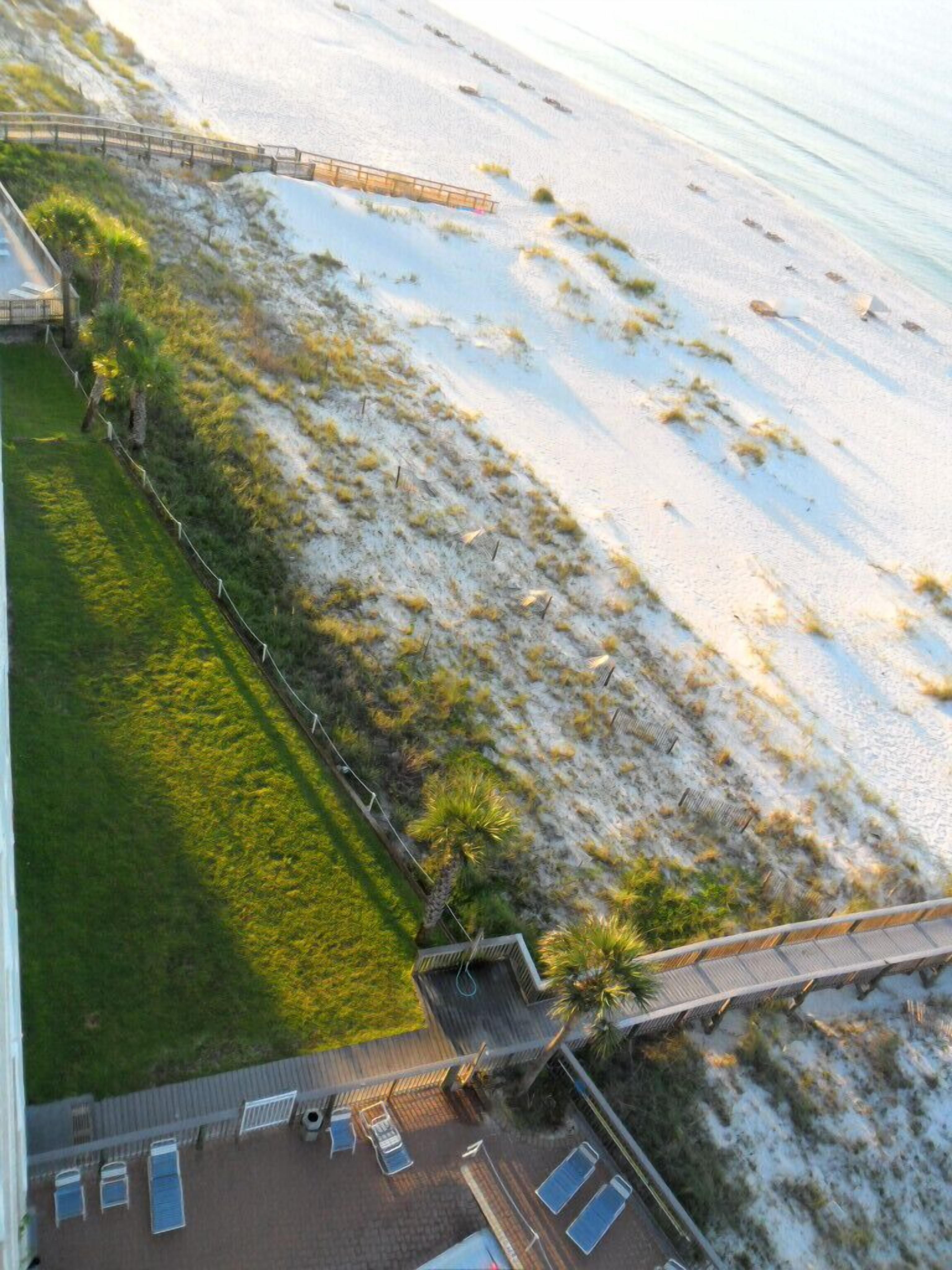 Vista aérea de um hotel à beira-mar, com caminho de acesso à praia, área gramada e estacionamento com espreguiçadeiras.