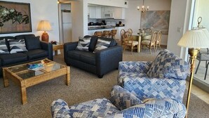 A living room with two sofas, a coffee table, and patterned cushions. There is a dining area with a table and chairs. The kitchen is visible in the background with white cabinets and appliances.
