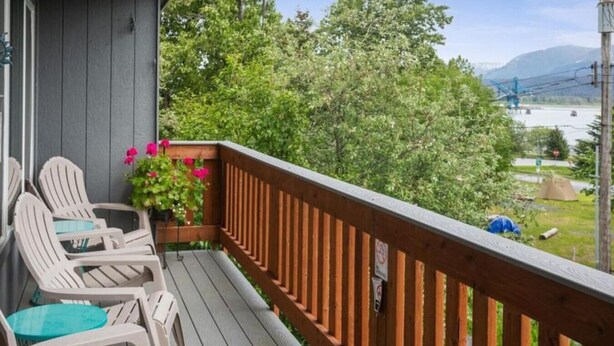 Una terraza de madera con sillas Adirondack y una mesa, con vista a un río y montañas.