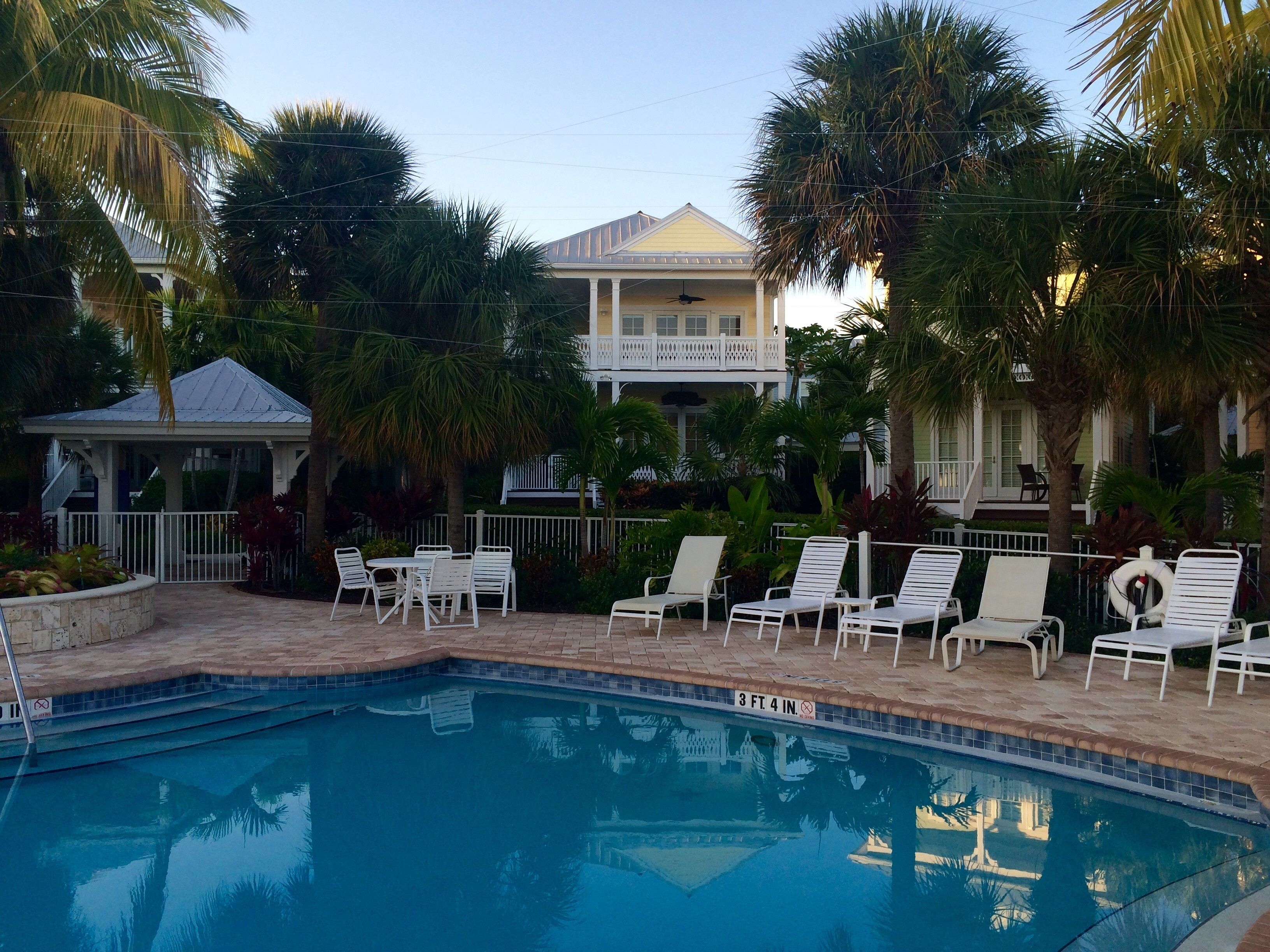 A pool area with lounge chairs, a pool with a depth marker, and a building with a balcony in the background.