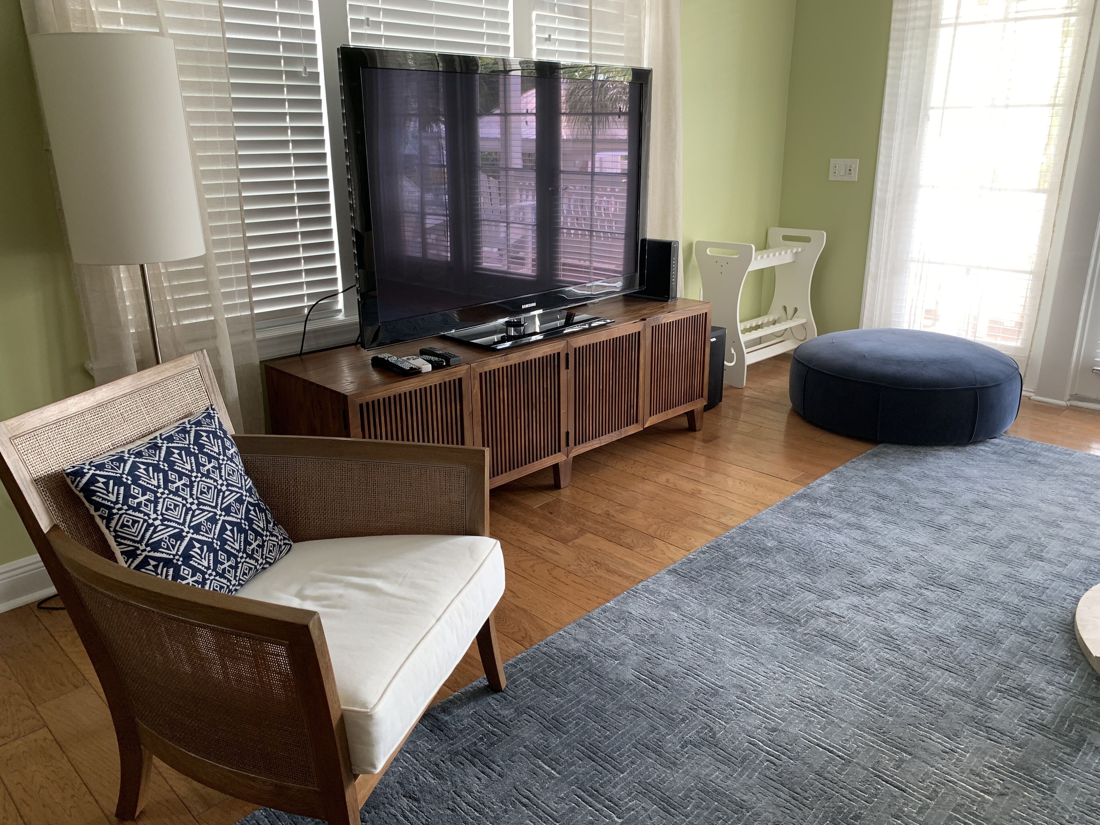 A living room with a television on a wooden stand, a blue patterned cushion on a wicker chair, a blue ottoman, and a grey rug.