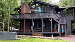 A two-story wooden cabin with a balcony, outdoor seating, and a covered patio area. - LAKESIDE ON LAKE NAOMI - boats, hot tub, beach, dock. Dogs OK (Pocono Pines)