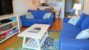 Ein Wohnzimmer mit blauem Sofa, einem weißen Couchtisch und einem Fernseher auf einem Schrank. Ein Bücherregal mit diversen Gegenständen und eine Küche sind im Hintergrund zu sehen.
