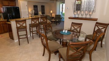A dining area with a glass table surrounded by wooden chairs, a kitchen in the background, and a ceiling fan.