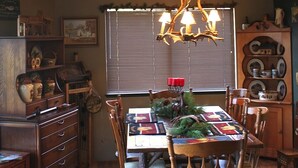 Un comedor con mesa de madera, sillas y una araña. Se aprecia una ventana con persianas, un aparador y diversos objetos decorativos en las paredes.