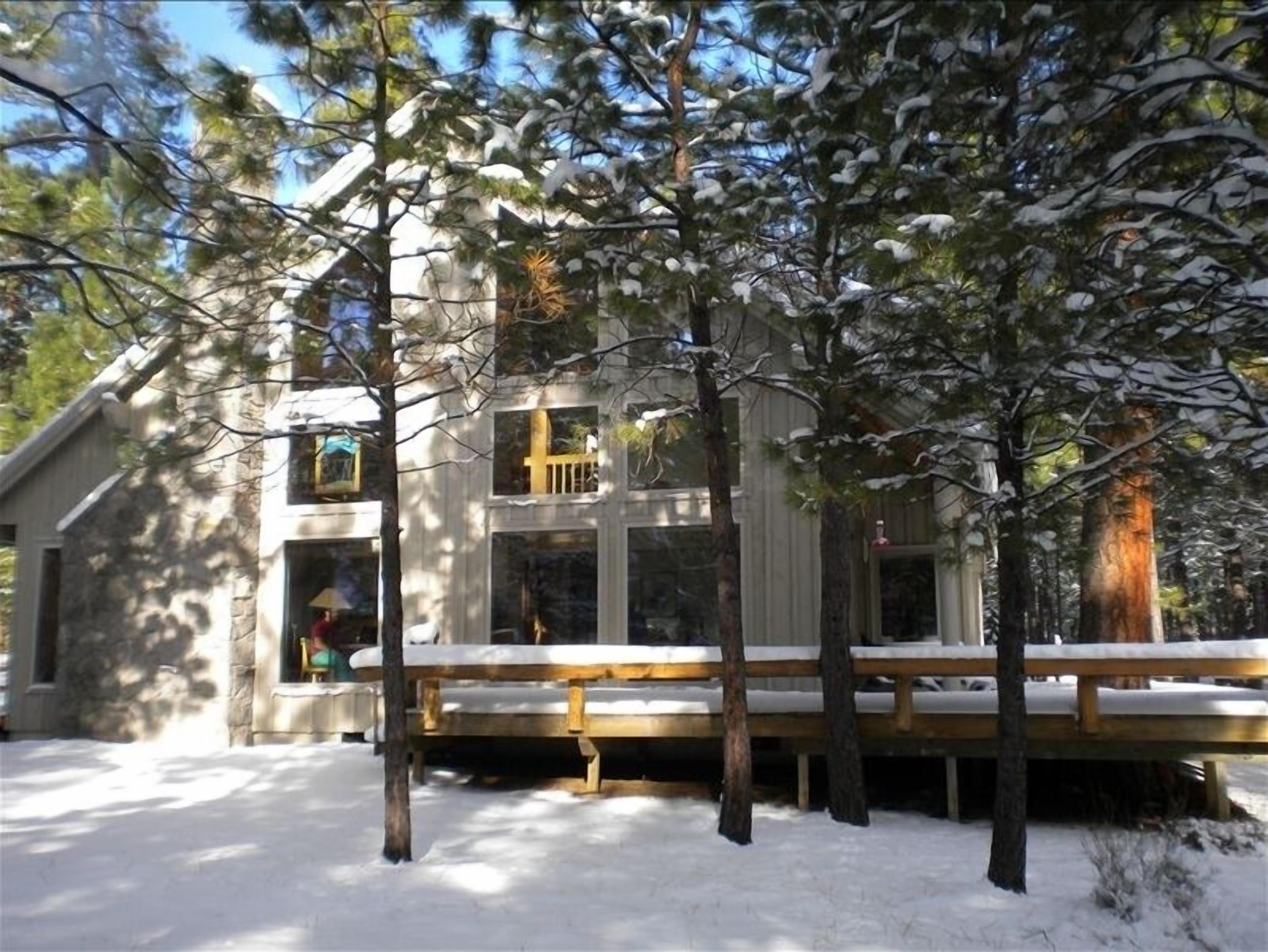 A two-story cabin with a wooden deck, surrounded by snow-covered trees.