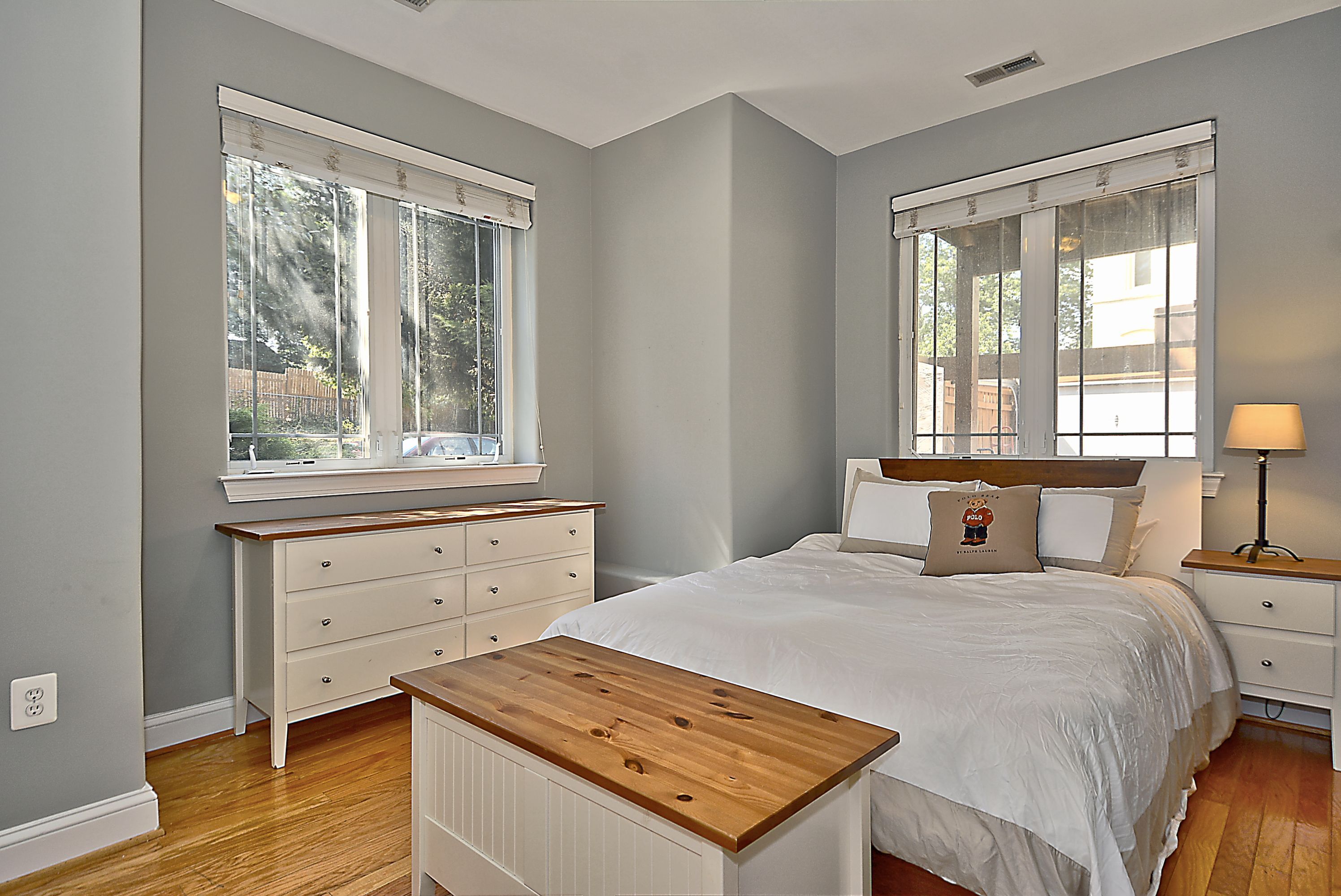 A bedroom with a wooden headboard, a white bed, a nightstand with a lamp, and a dresser with drawers.