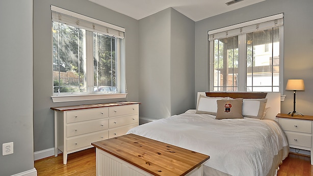 Un dormitorio con cabecera de madera, cama blanca, mesita de noche con lámpara y cómoda con cajones.