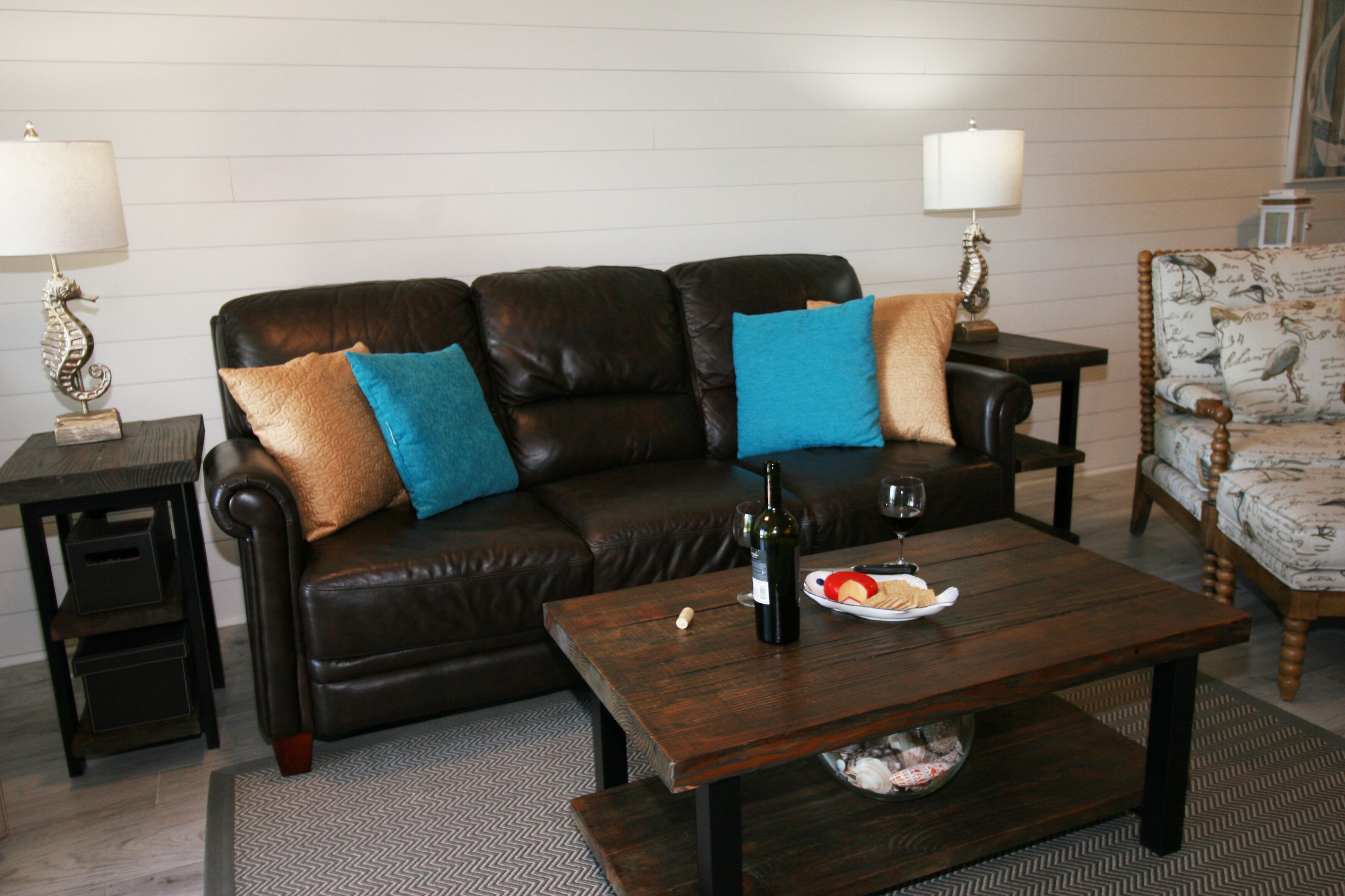 A living room with a dark brown leather sofa, two side tables with lamps, a wooden coffee table with a bottle and plate of food, and a patterned armchair.