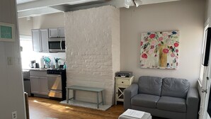 Ett kompakt bostadsutrymme med ett litet kök, en grå soffa, ett litet bord och en säng med ett sängöverkast med marint tema.