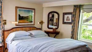 A neatly made bed with a striped bedspread and pillows, a framed landscape painting above it, a mirror on the wall, and a window with curtains. - Garden Apartment Located in a Wonderful Old Victorian Home (Alameda)