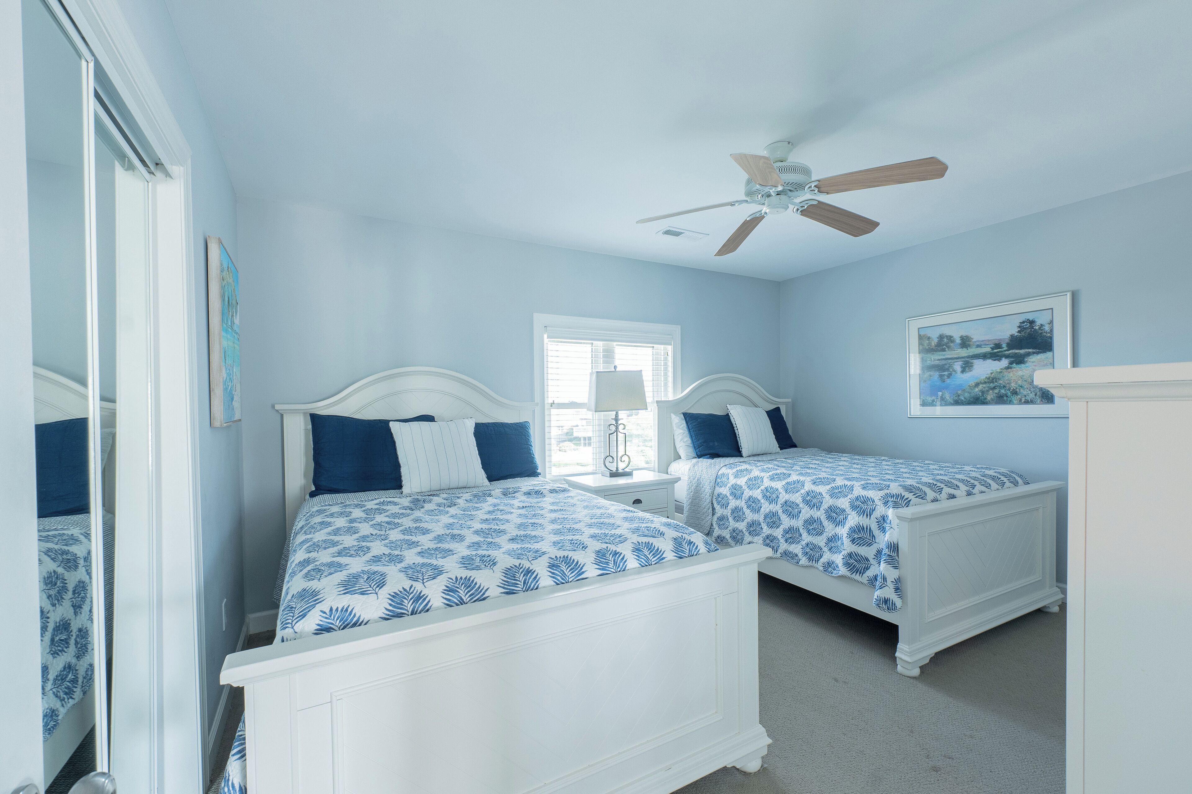 A bedroom with two beds, a ceiling fan, a window, and a painting on the wall.