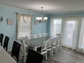 Une salle à manger avec une table rectangulaire entourée de chaises blanches, un lustre et deux fenêtres ornées de rideaux légers.