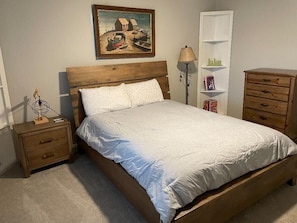 A bedroom with a wooden bed, a nightstand with drawers, a lamp, a bookshelf, and a painting on the wall.