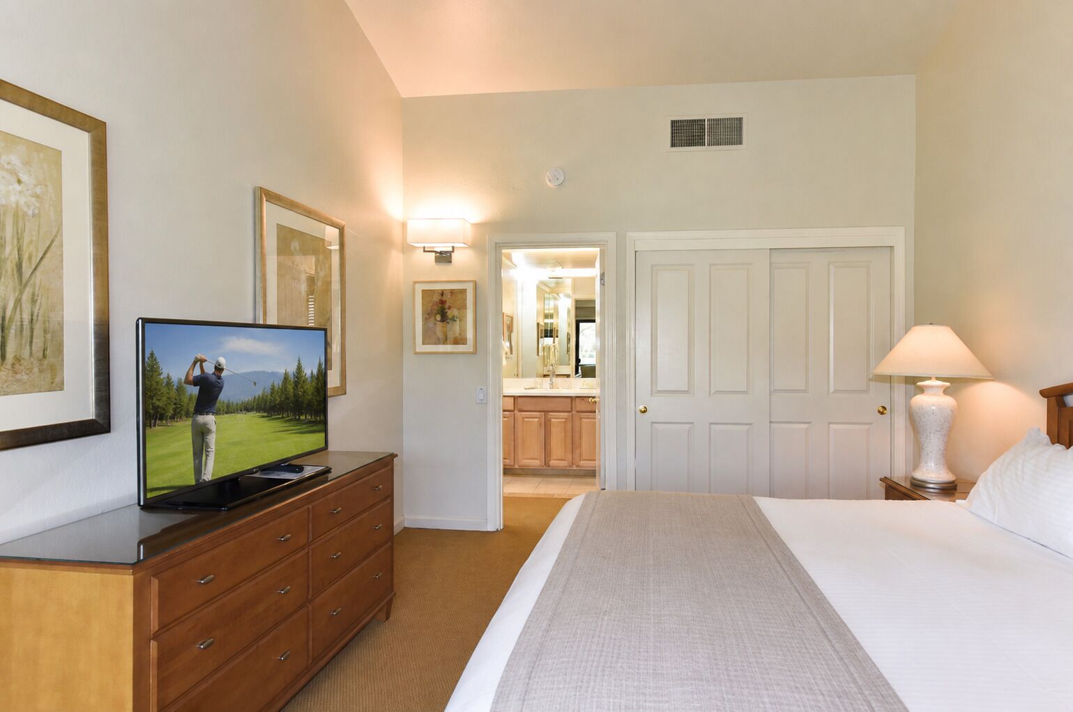 A hotel room with a bed, a television, a dresser, a lamp, and a door leading to a bathroom.