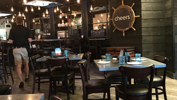 L’intérieur d’un restaurant, avec des tables, des chaises, des ampoules suspendues et un panneau sur lequel on peut lire « À votre santé ! ».