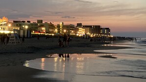 Une plage avec du sable mouillé et des reflets de bâtiments. On y voit des gens qui se promènent et qui sont assis sur le sable.
