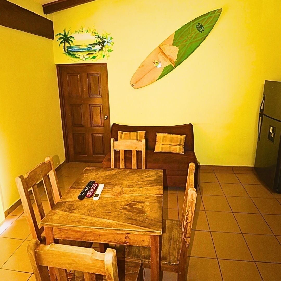 A dining area with a wooden table and chairs, a sofa with cushions, a wall-mounted surfboard, and a door.