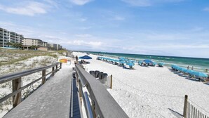 A beach with a wooden boardwalk leading to the shore, lined with blue umbrellas and lounge chairs.