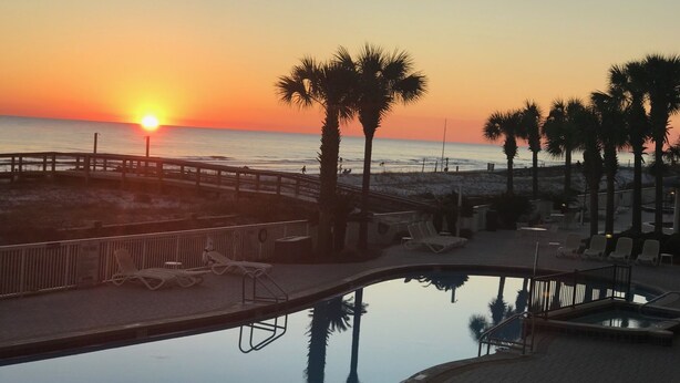 A pool area with lounge chairs, a wooden deck, and a view of the sunset over the ocean.