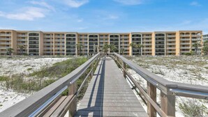 A wooden boardwalk leading to a multi-story hotel with balconies, situated on a sandy beach.