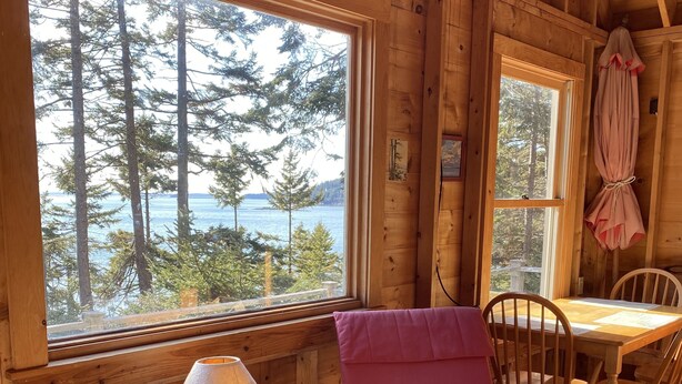 En mysig stuga med en stor fönsteröppning som ger utsikt över skogen och vattnet. Ett träbord och stolar står placerade nära fönstret, och en lampa står på ett litet bord bredvid stolen.