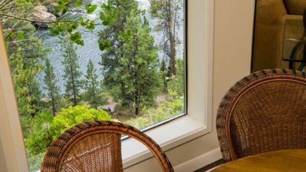 Comedor con sillas de mimbre y una mesa lista para dos, con vista a un río y bosque a través de un ventanal grande.