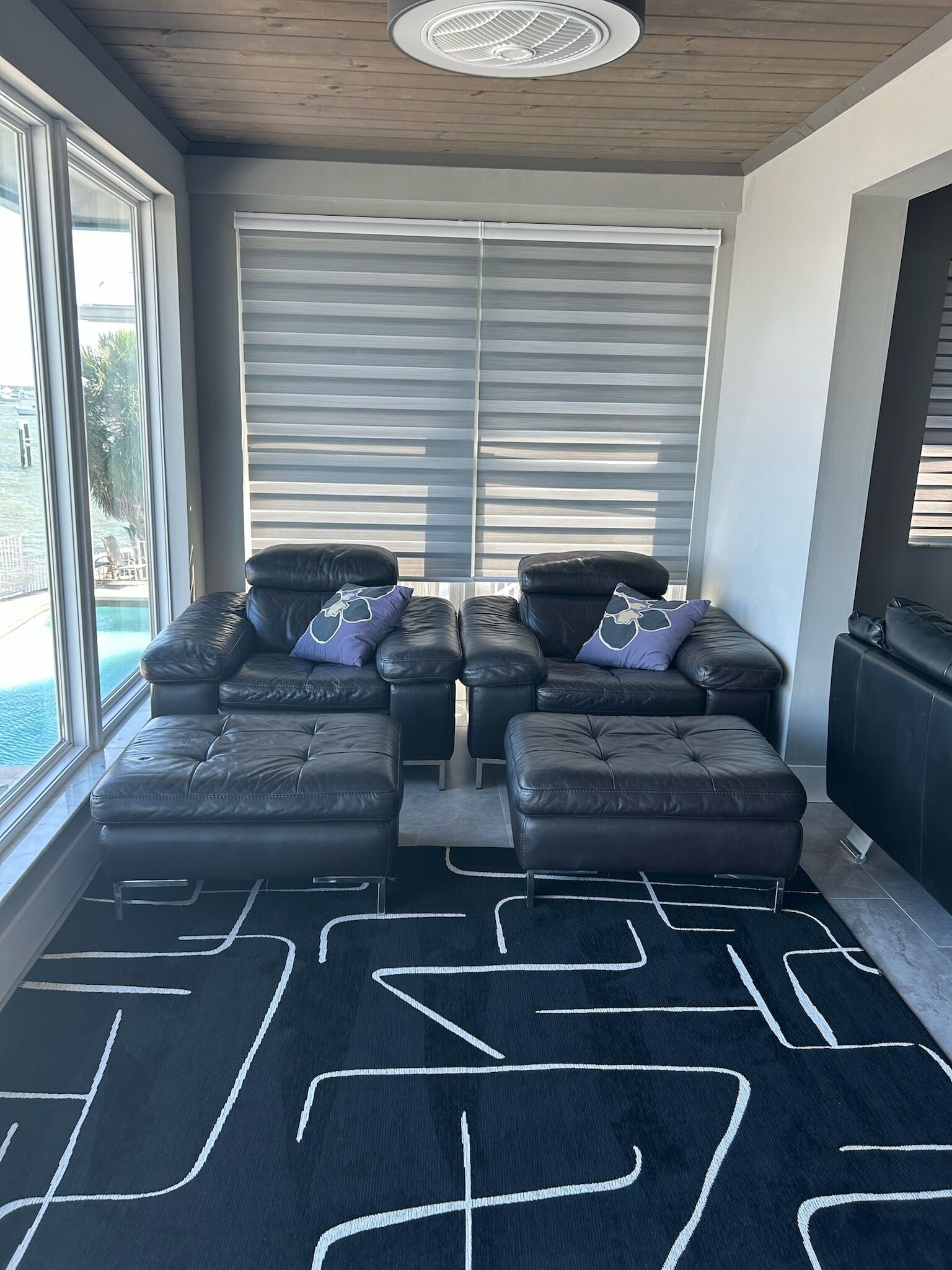 A modern living room with a black leather sofa set, a large window with blinds, and a view of a pool.