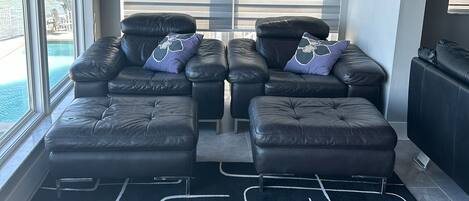 A modern living room with a black leather sofa set, a large window with blinds, and a view of a pool.