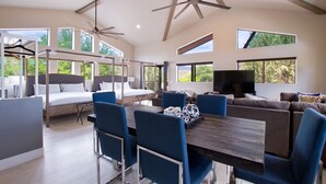 Een ruime kamer met een grote eettafel, blauwe stoelen, een bank en een televisie. Ramen zorgen voor natuurlijk licht en uitzicht op groen.